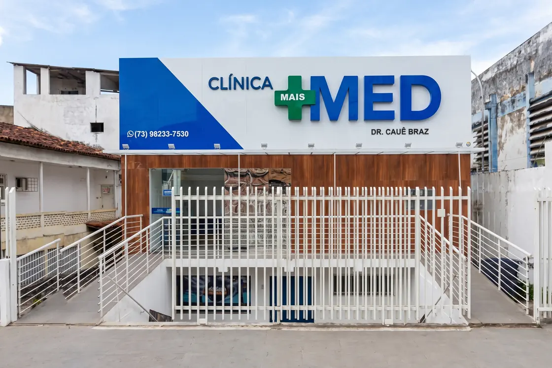 Fachada da Clínica Mais Med, com a placa branca e azul sobre o gradil da entrada